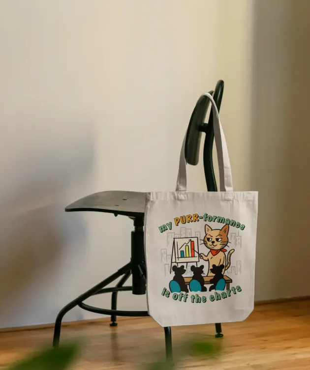 Geantă tote ecru cu print pisică și mesajul „my PURR-formance is off the charts”, fotografiată pe un scaun.