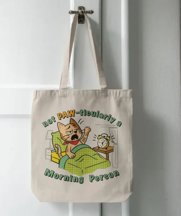 Geantă tote ecru cu print pisică în pat și mesajul „Not PAW-ticularly a Morning Person”, agățată pe ușă.
